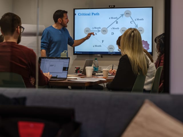 Team meeting at Murmur Creative discussing project timelines, with a presenter pointing at a chart labeled "Critical Path" on a screen.